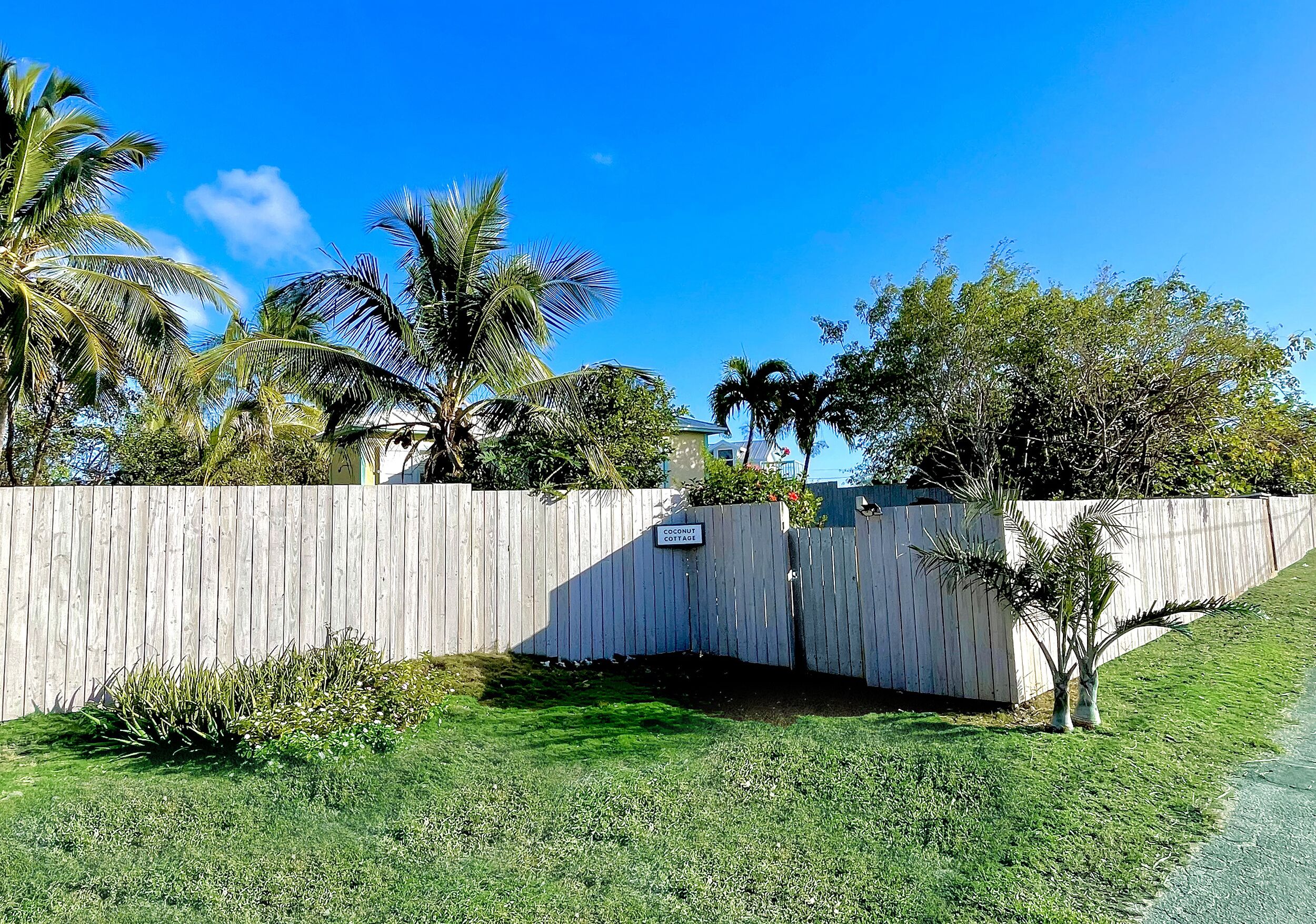 Ein umzäunter Garten mit einem Schild, das "Coconut Cottage" zeigt, Palmen und einem strahlend blauen Himmel.