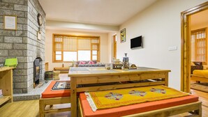 A cozy room with a stone fireplace, a wooden table with a yellow mat, a sofa, and a TV on the wall. - GuestHouser 1 BR Cottage 4e68 (Manali)