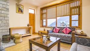 A cozy living room with a sofa, armchairs, a coffee table, and a fireplace. There is a large window with a view of mountains and a wooden door. - GuestHouser 1 BR Cottage 4e68 (Manali)