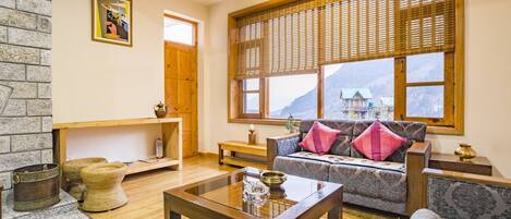 A cozy living room with a sofa, armchairs, a coffee table, and a fireplace. There is a large window with a view of mountains and a wooden door.