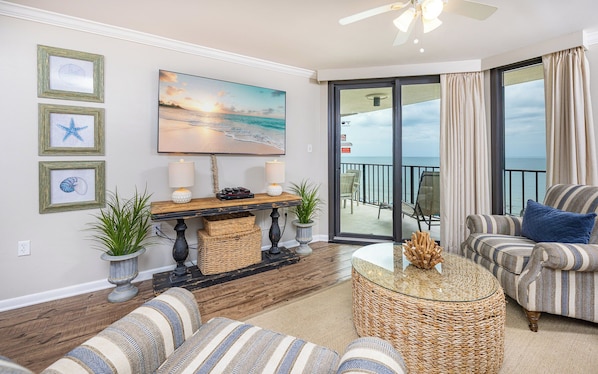 A living room with a view of the ocean, a ceiling fan, and a large painting.