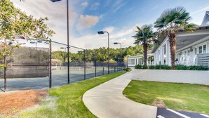 A tennis court with a fence, a pathway, and a building with a white exterior and palm trees. - Legasea DeBordieu Home LegaSea Spectacular porches Close to Beach (Georgetown)