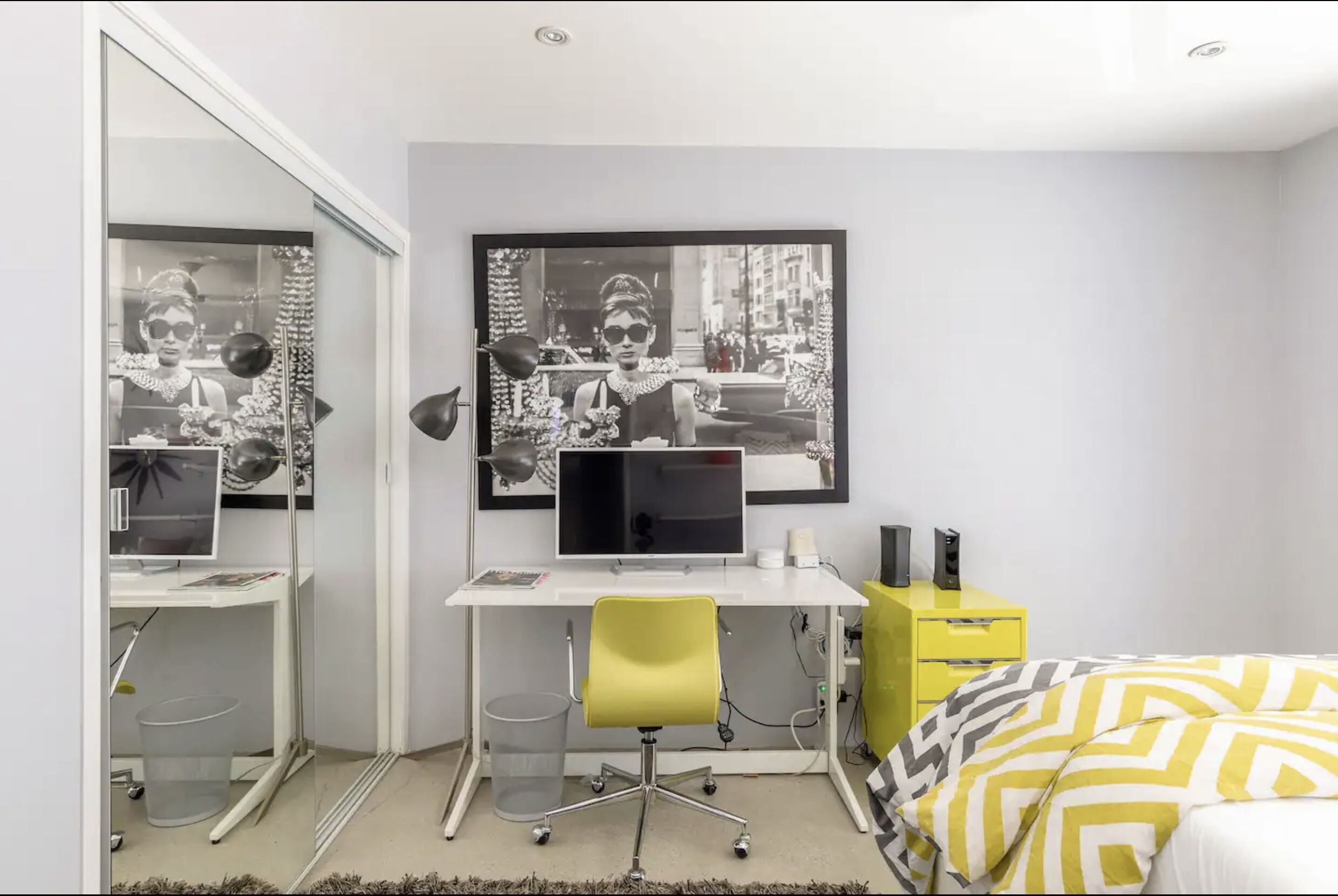 A modern bedroom with a white desk, a yellow chair, a bed with patterned bedding, and two black-and-white framed pictures on the wall.