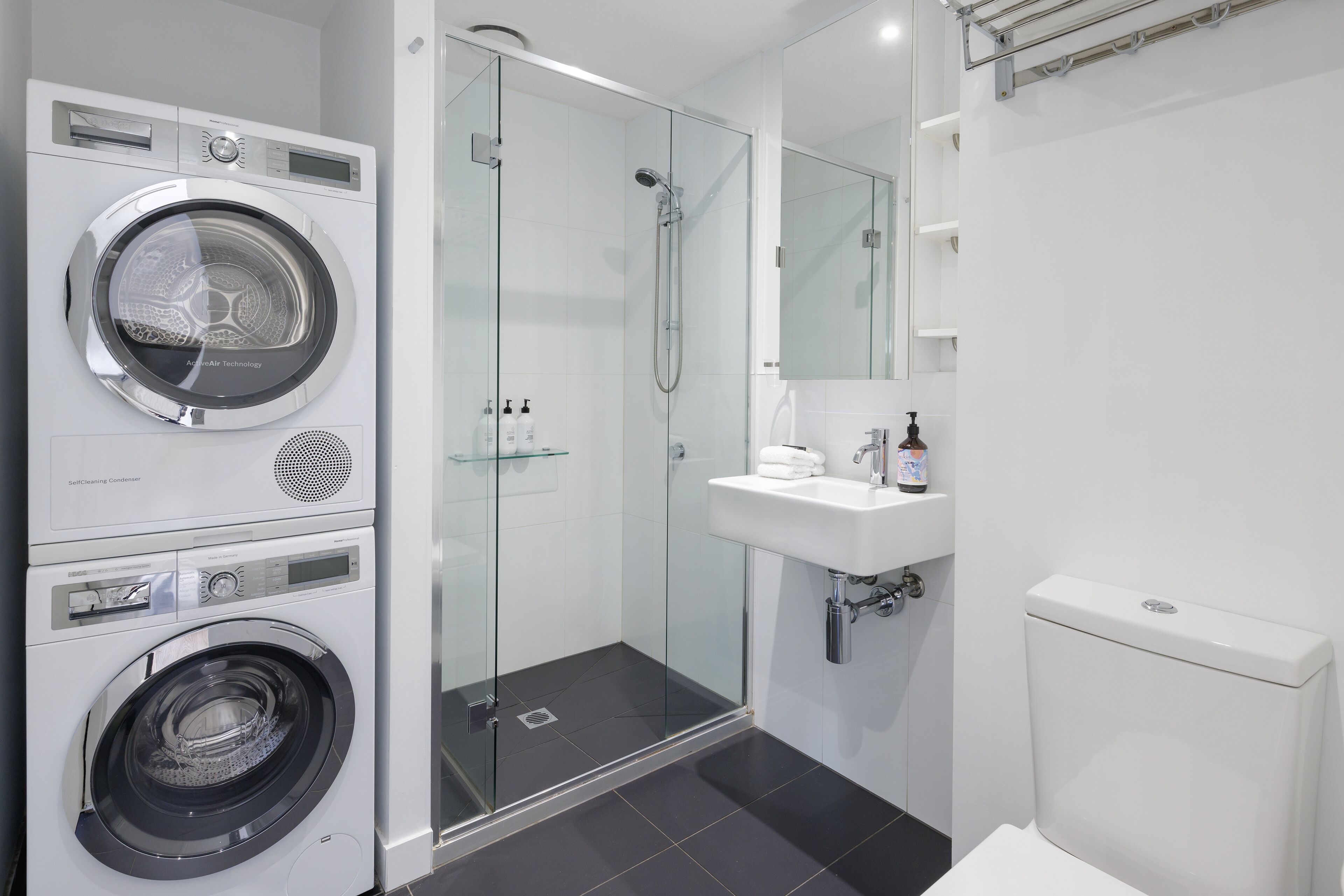A compact hotel bathroom with a washing machine, a glass-enclosed shower, and a sink with a mirror above it.