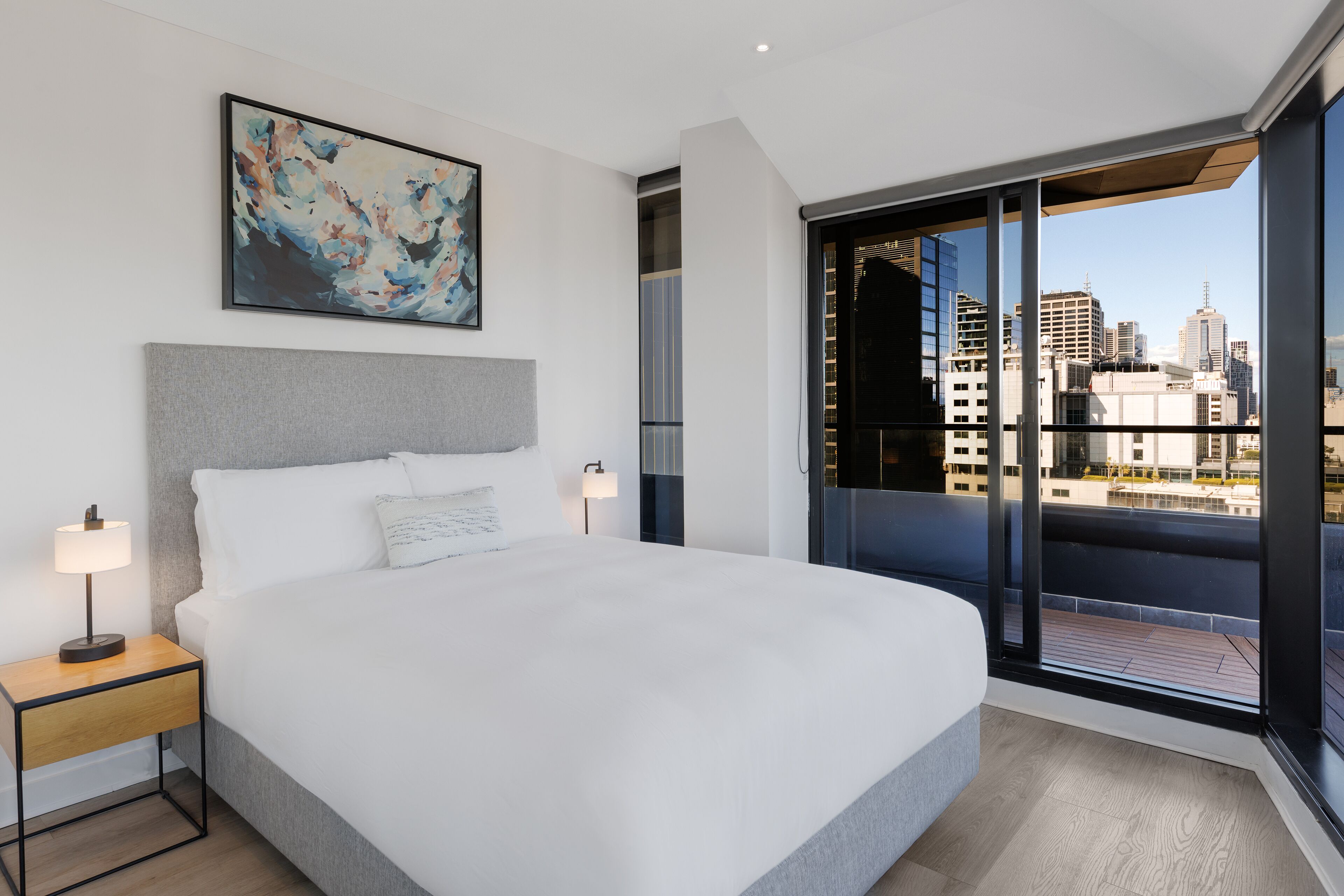 A modern hotel room with a large bed, a city view balcony, and a painting on the wall.