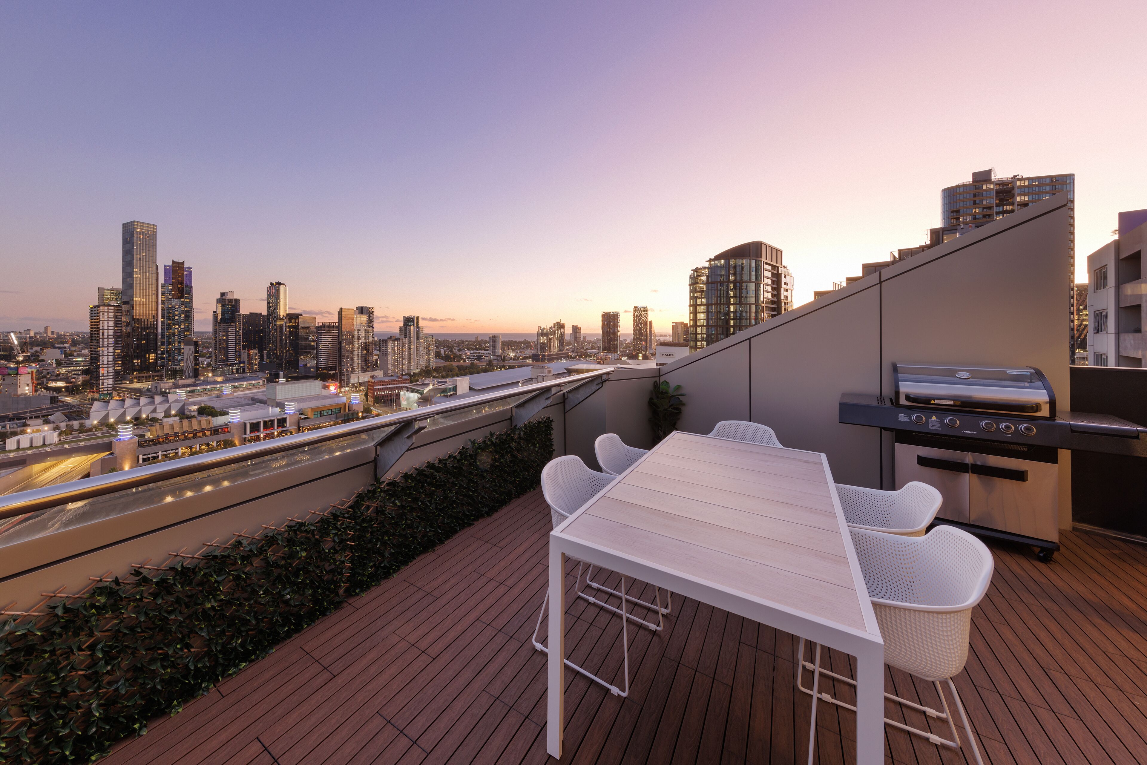 A rooftop terrace with a dining table and chairs, a grill, and a view of a city skyline at dusk.