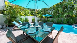 Uma mesa de jantar com tampo de vidro, cadeiras, um guarda-sol azul e uma piscina ao fundo.