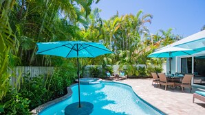Piscina com design curvo, rodeada por vegetação exuberante e palmeiras. Há dois guarda-sóis oferecendo sombra e uma área de refeições com cadeiras e mesa.