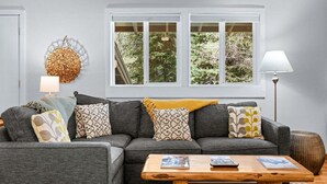 A grey sectional sofa with patterned cushions, a wooden coffee table, a wall-mounted round decorative item, and a window with a view of greenery. - RMR: Gros Ventre A5 Condo in Teton Village (Teton Village)