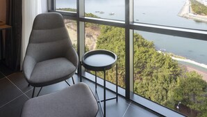 Una habitación moderna con ventanas amplias, dos sillas grises, una mesita redonda y vistas al agua y vegetación.