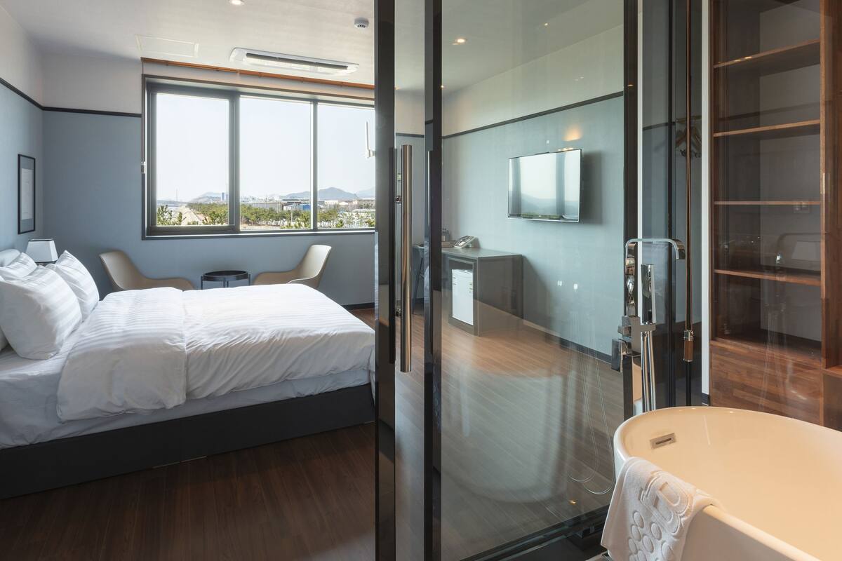 A modern bedroom with a large bed, a glass door leading to a bathroom, and a city view.