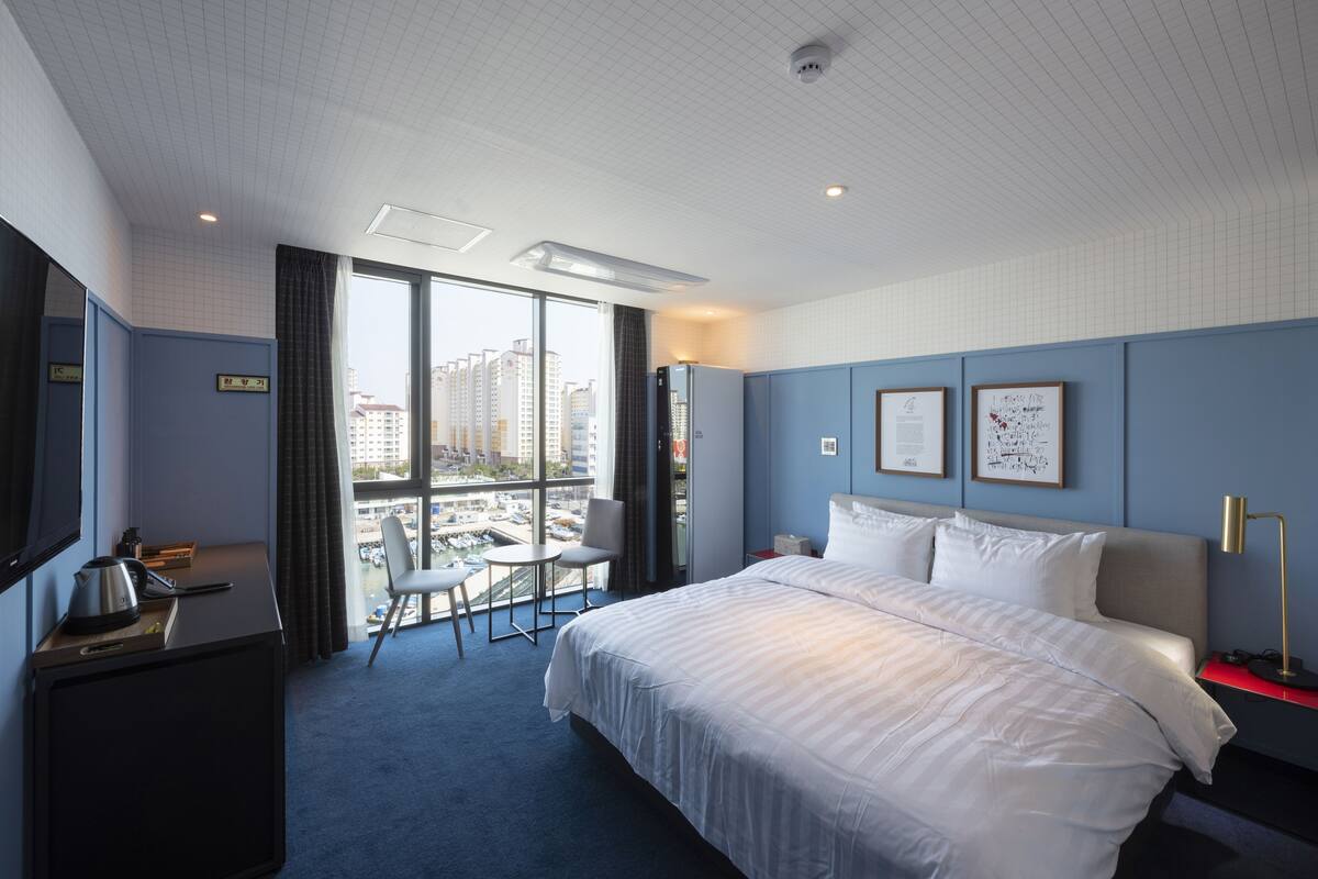 A hotel room with a large bed, a desk with a kettle, a chair, and a view of the city through large windows.