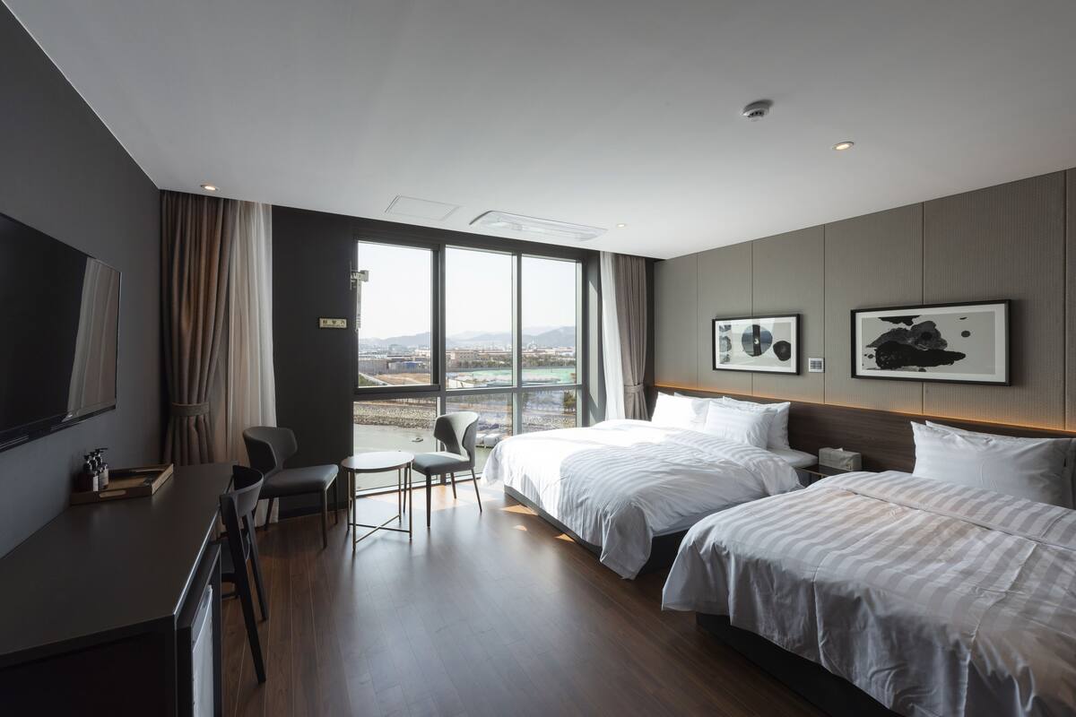 A modern hotel room with two beds, a desk, and a view of the city.
