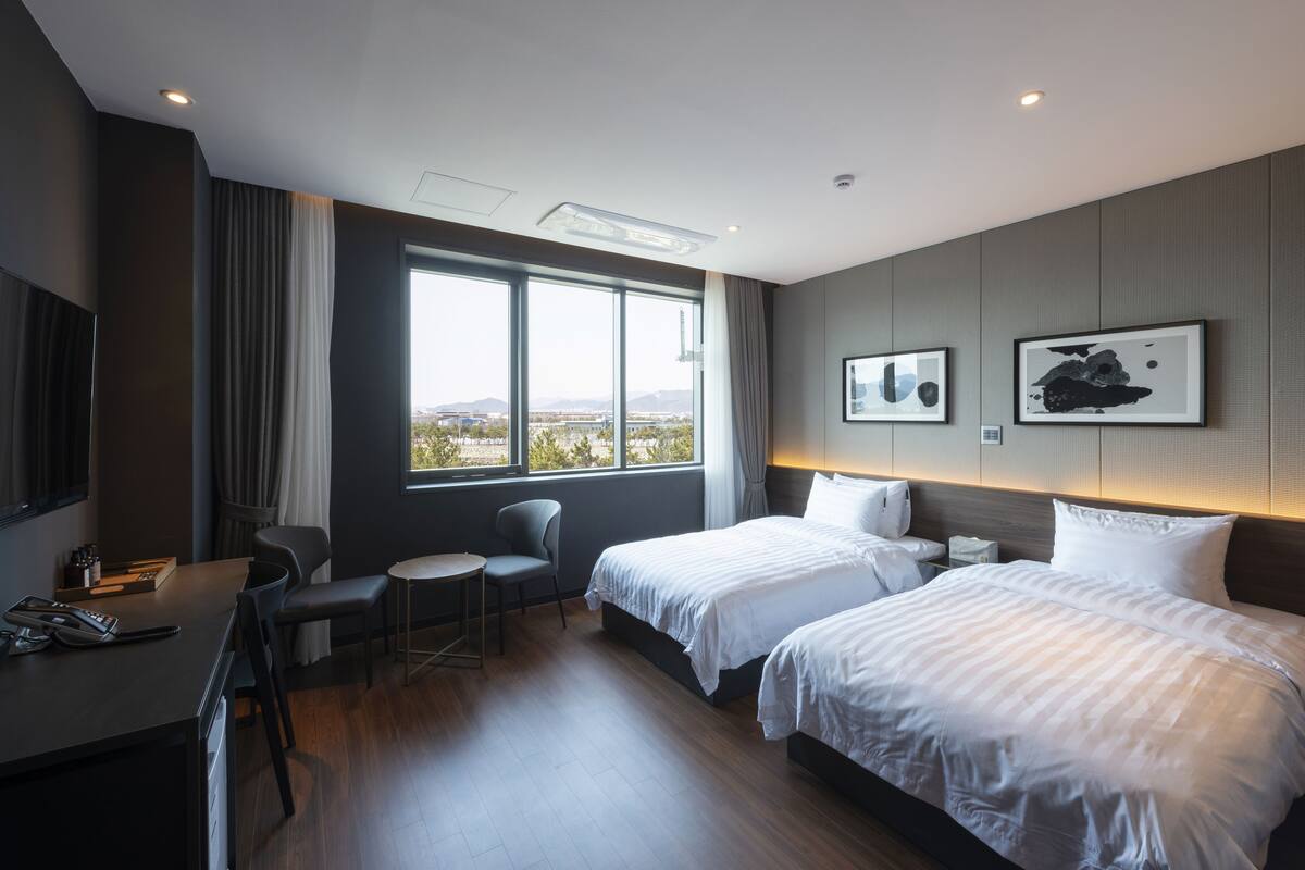 A hotel room with two beds, a desk, a chair, and a large window offering a view of the outdoors.