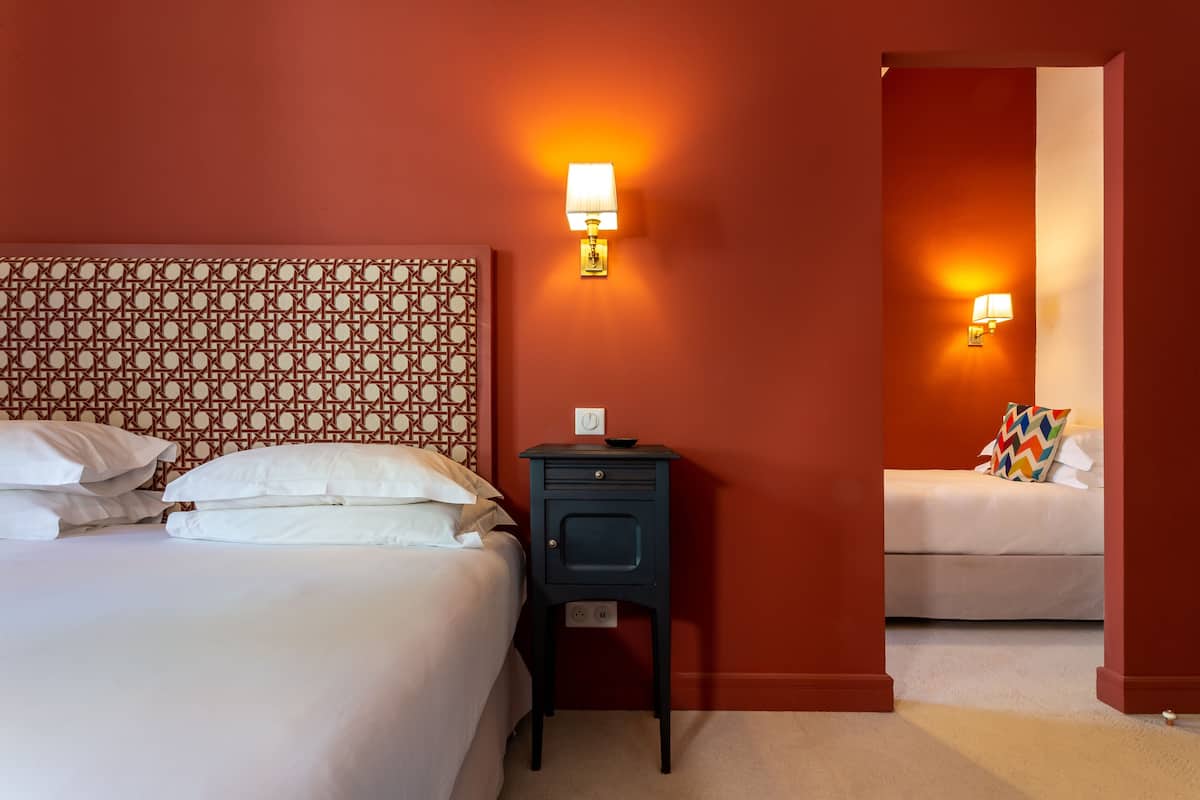 Une chambre d’hôtel avec une tête de lit à motifs, deux tables de chevet avec des lampes, un lit avec du linge de lit blanc et une vue sur la pièce adjacente.
