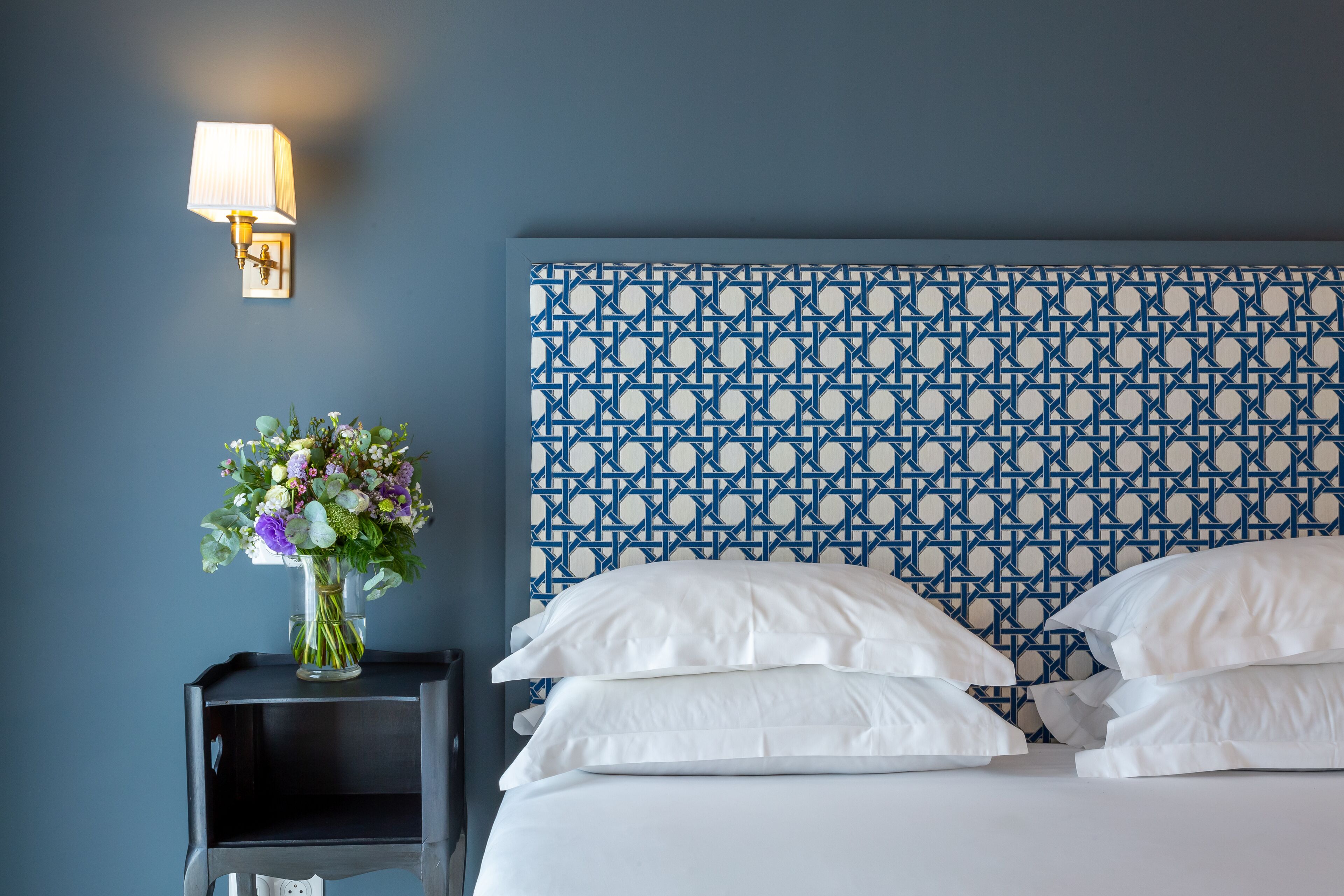 A neatly made bed with white linens, a wall-mounted light fixture, and a patterned blue and white headboard.