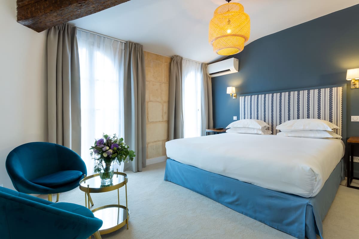 Une chambre d’hôtel avec un grand lit, deux chaises bleues, une petite table ronde ornée d’un vase de fleurs et un luminaire suspendu.
