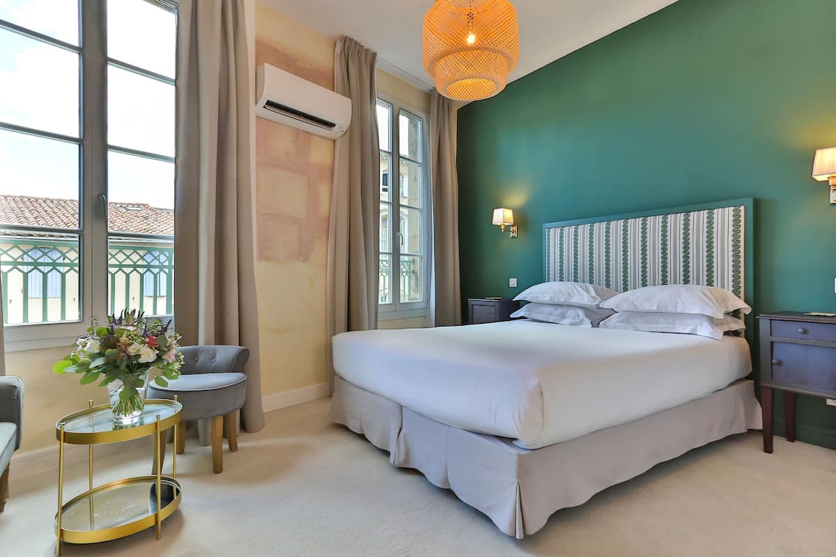 Une chambre d’hôtel avec un grand lit, une table ronde en verre ornée d’un vase de fleurs, un fauteuil gris et une vue sur le balcon extérieur.