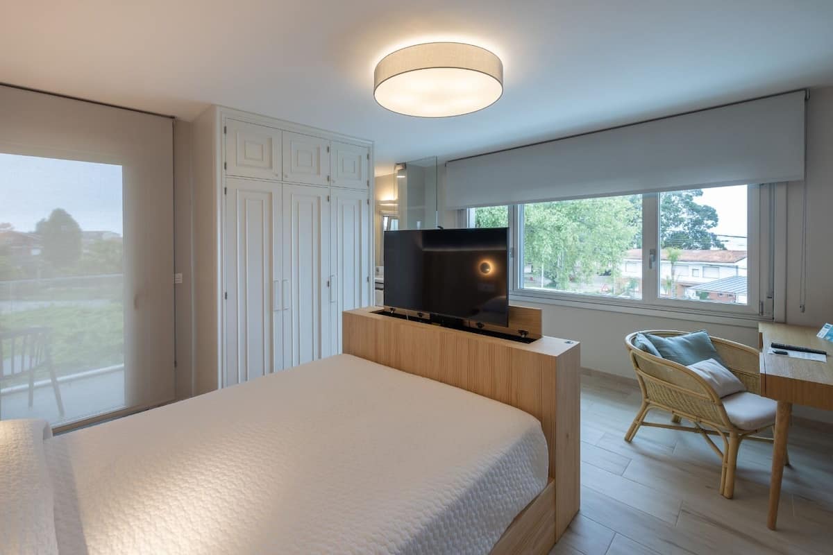 Quarto moderno com cama, televisão em suporte de madeira, escrivaninha com cadeira e janelas amplas.