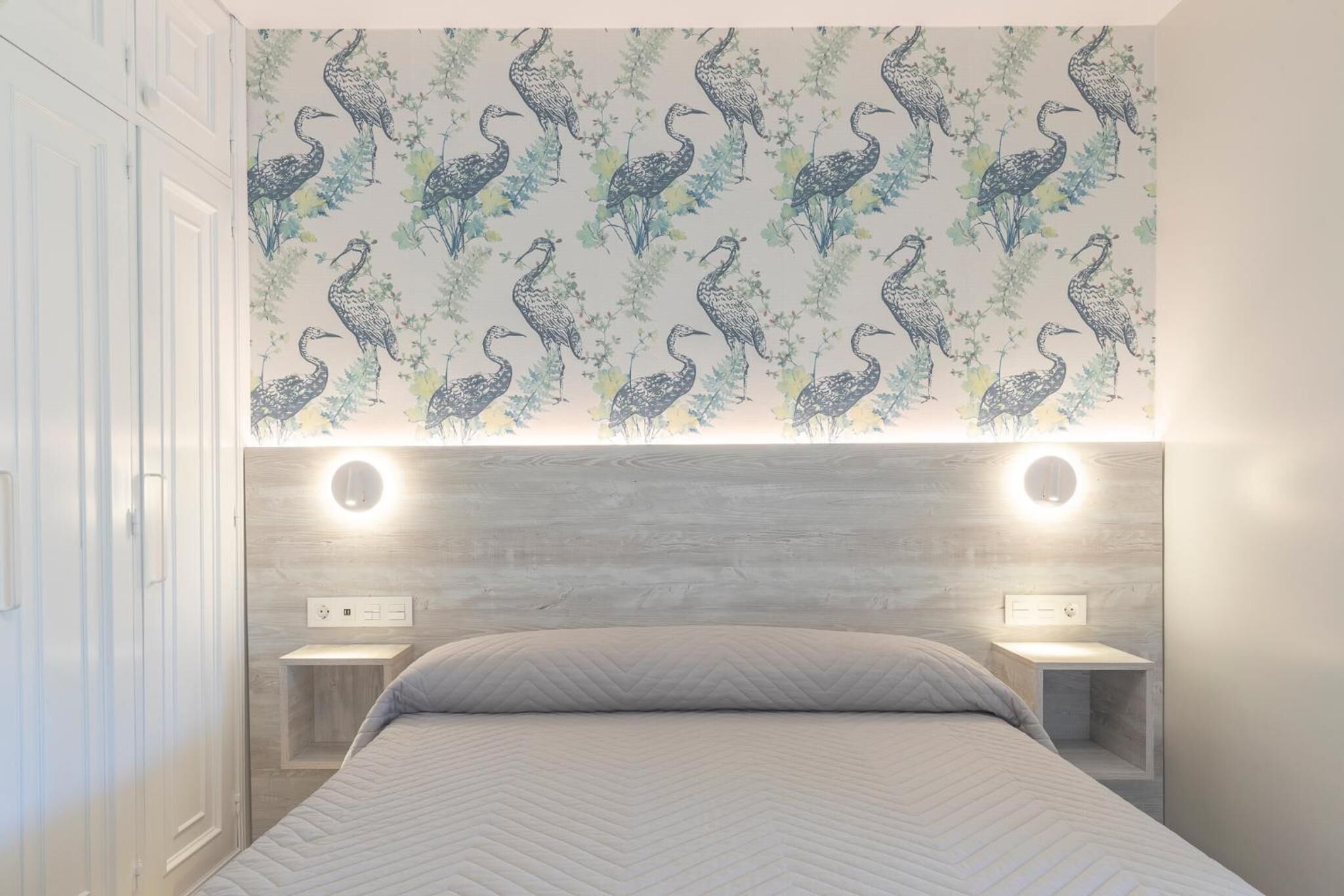A bedroom with a patterned wallpaper, a bed with a herringbone quilt, and bedside tables with built-in lighting.