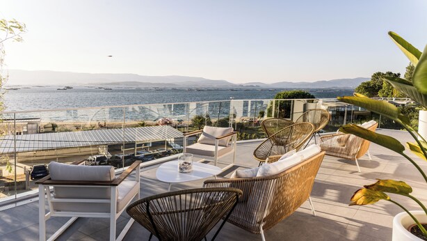 A terrace with wicker furniture, a glass balustrade, and a view of the sea and distant mountains.