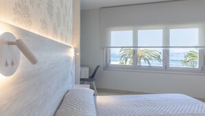 A bedroom with a large bed, a desk, and a window with a view of palm trees and the sea.