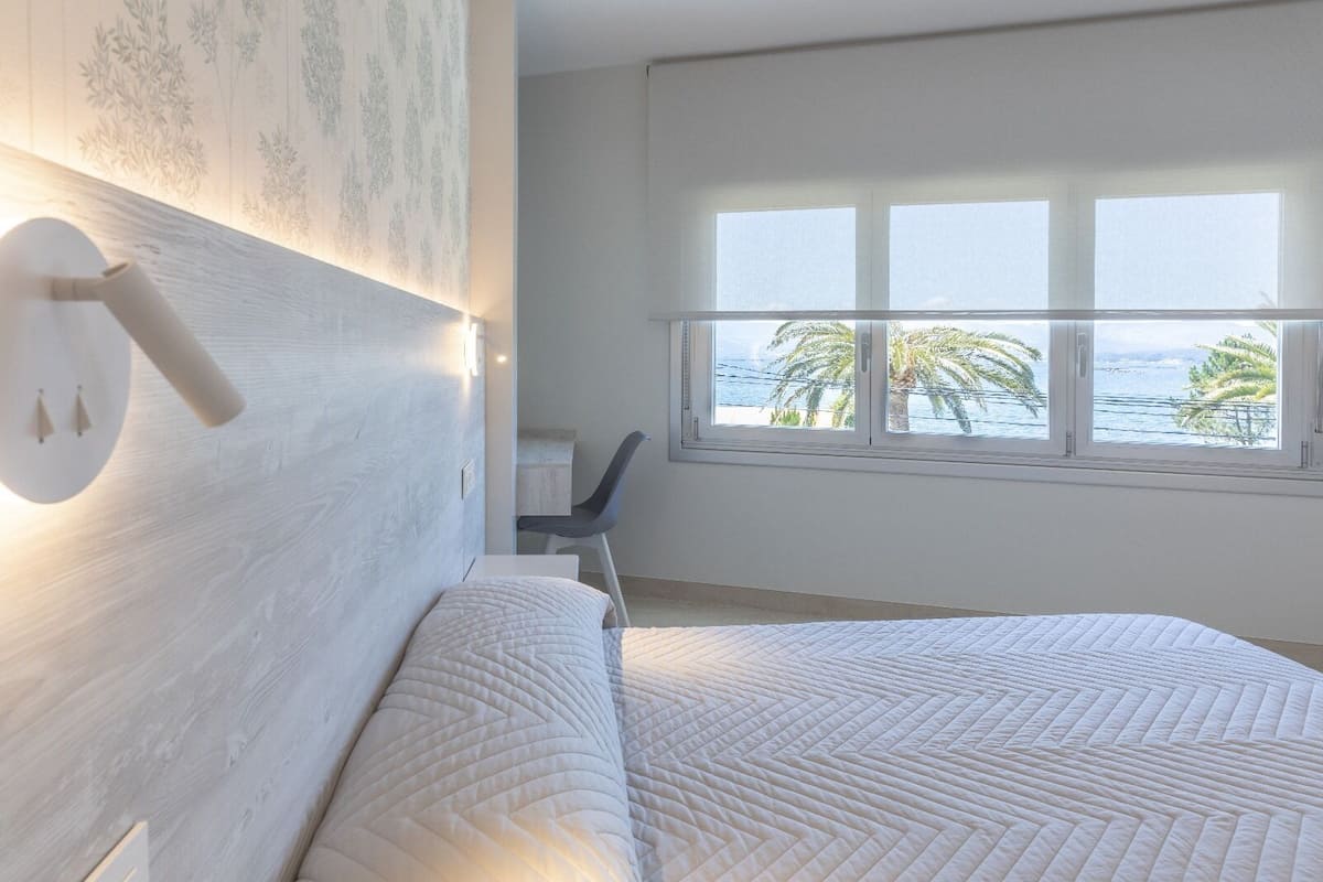Quarto com cama grande, escrivaninha e janela vista para palmeiras e mar.