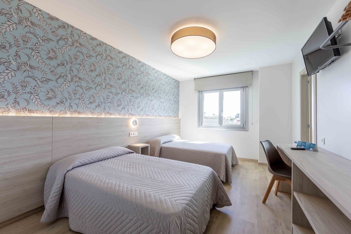 Quarto de hotel moderno com duas camas, uma escrivaninha, televisão e janela.