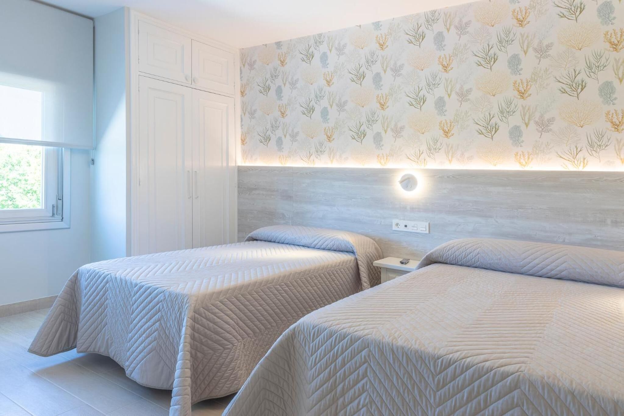 Een slaapkamer met twee bedden, een raam en een muur met een bladpatroon.