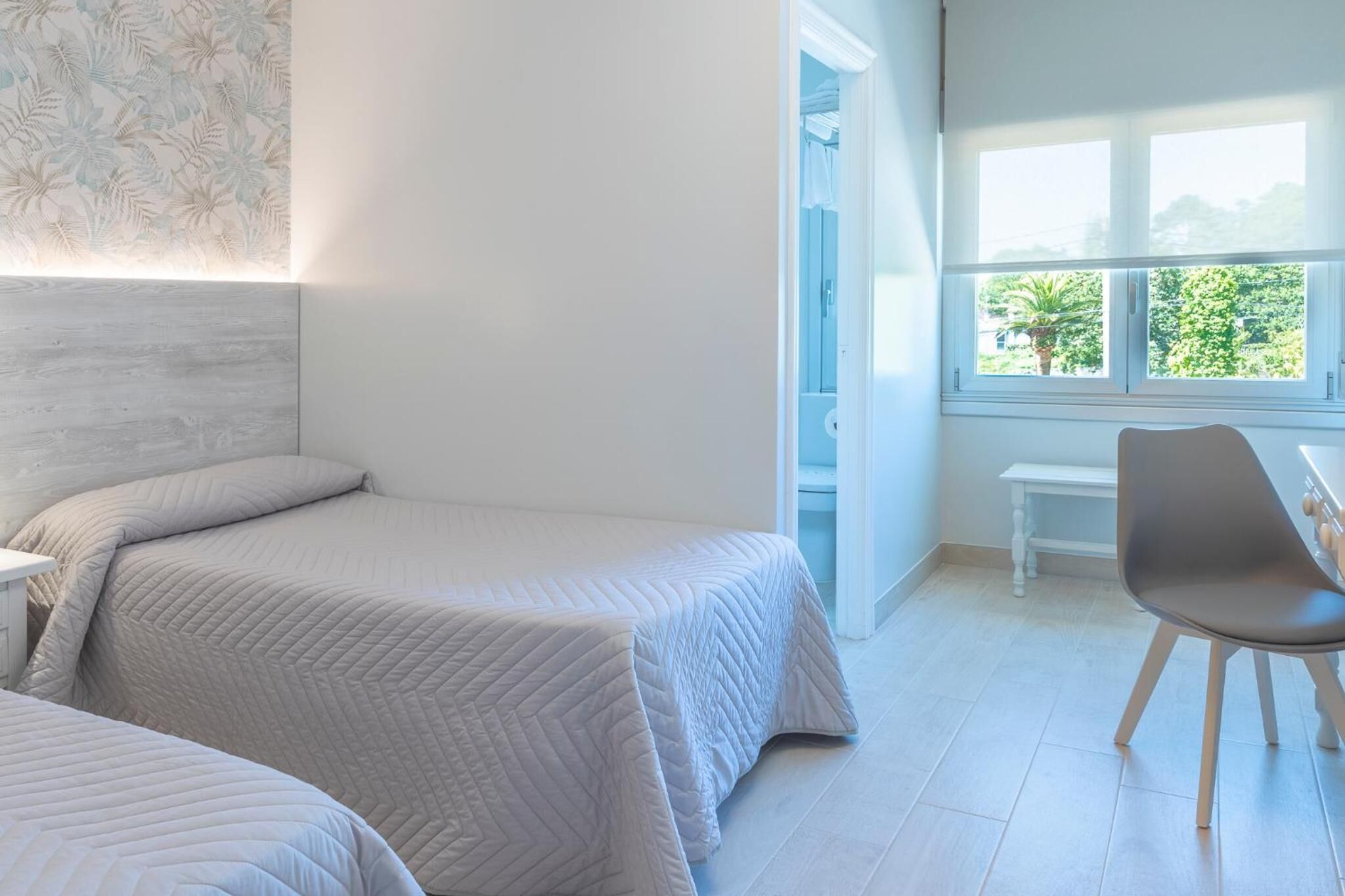 A bedroom with two beds, a chair, a small table, and a window with a view of greenery.