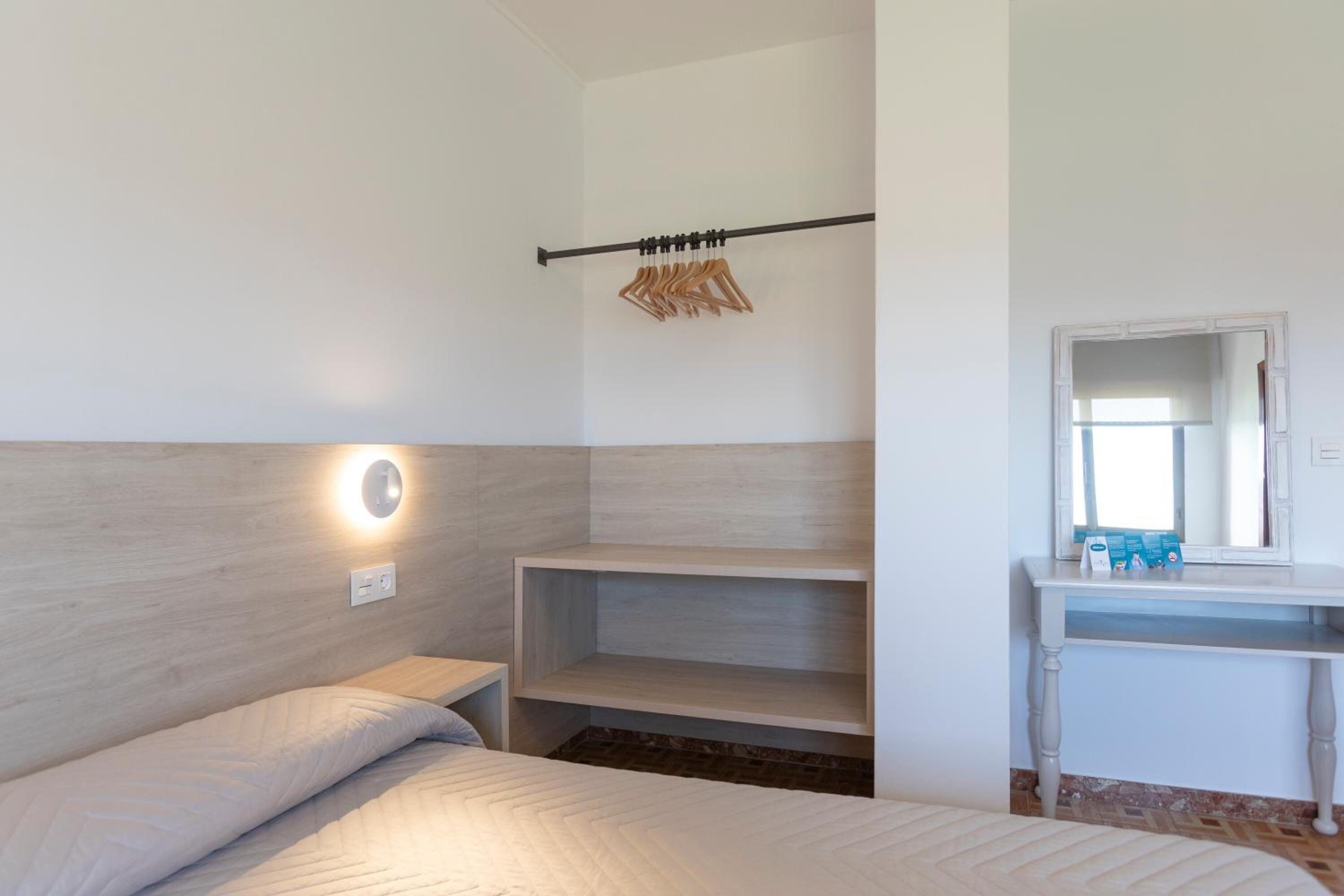 A bedroom with a bed, a bedside table, a wall-mounted shelf, a mirror, and a clothing rack.