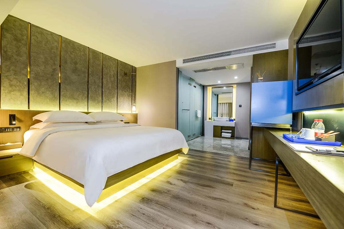 A modern hotel room with a large bed, a desk with a flat-screen TV, and a bathroom visible through an open door.