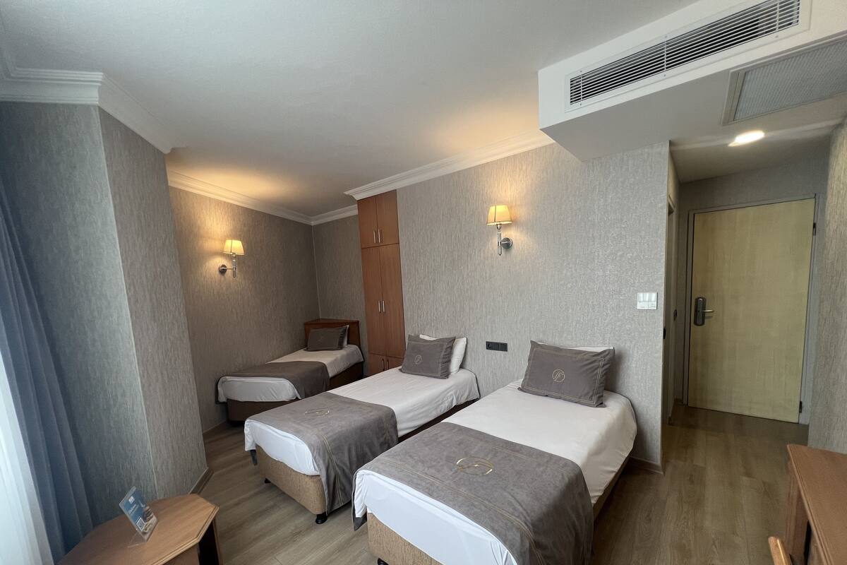 Ein Hotelzimmer mit drei Einzelbetten, einem Holzschrank, einem kleinen Holztisch und einer Wandlampe.