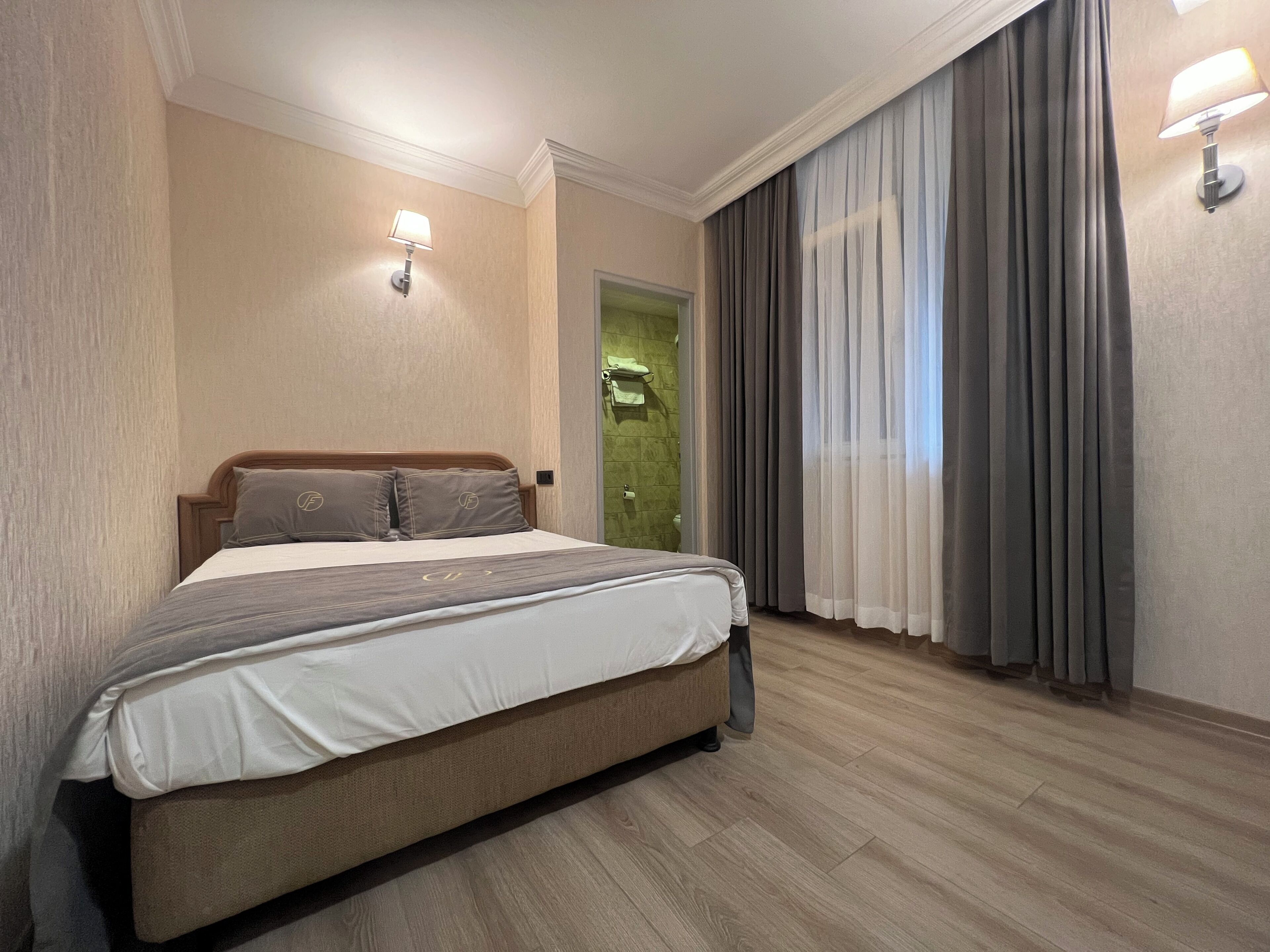 Habitación de hotel con una cama grande, cabecera de madera, puerta verde y ventana con cortinas grises.