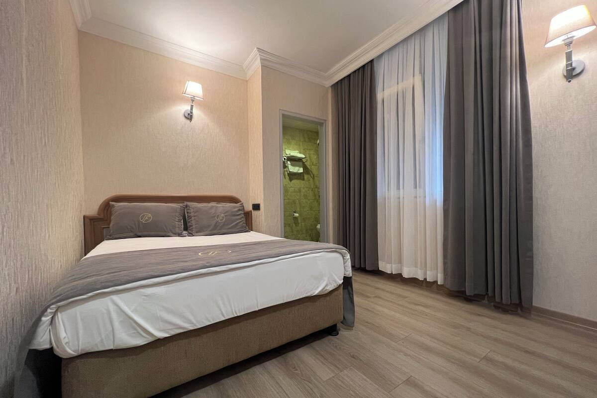 Habitación de hotel con una cama grande, cabecera de madera, puerta verde y ventana con cortinas grises.