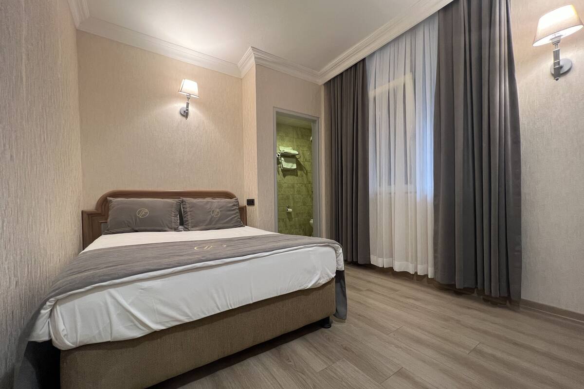 Ein Hotelzimmer mit einem großen Bett, einem hölzernen Kopfteil, einer grünen Tür und einem Fenster mit grauen Vorhängen.