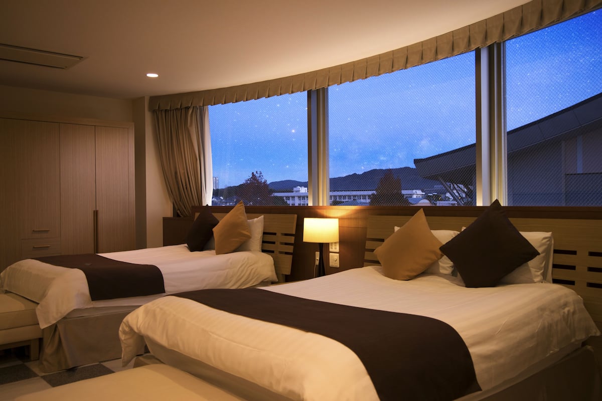 A hotel room with two beds, a nightstand with a lamp, and a large window offering a view of the cityscape.