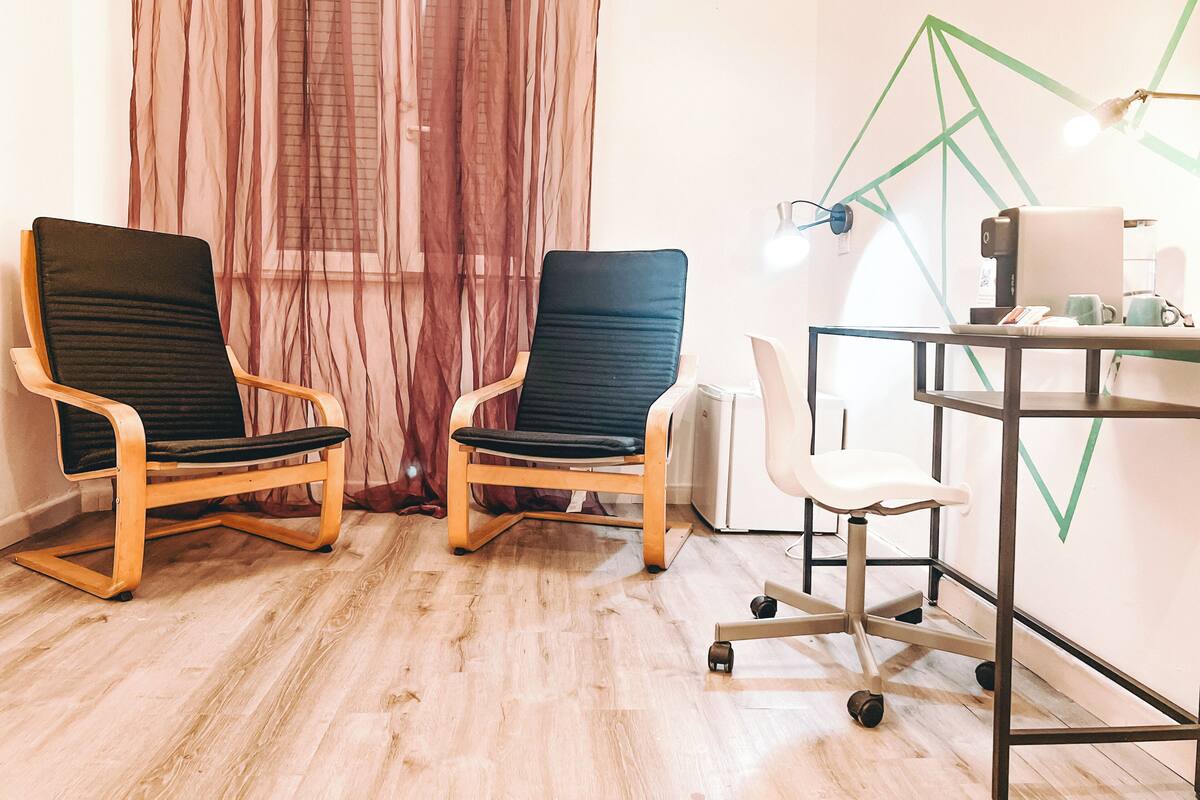 Una habitación con dos sillas negras, una silla blanca, un escritorio con una computadora y una pared con diseños geométricos.