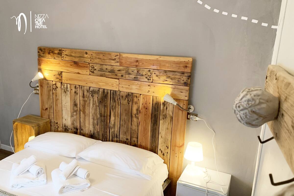 Un dormitorio con cabecera de madera, dos lámparas de noche, una mesita de noche y toallas dobladas con cuidado.