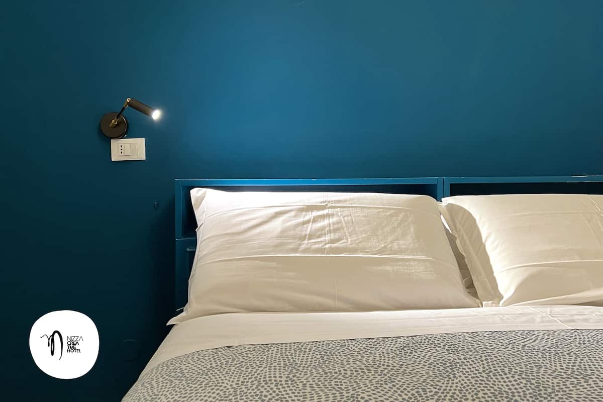 Una cama con ropa blanca, un cabecero azul y una luz nocturna en la pared.