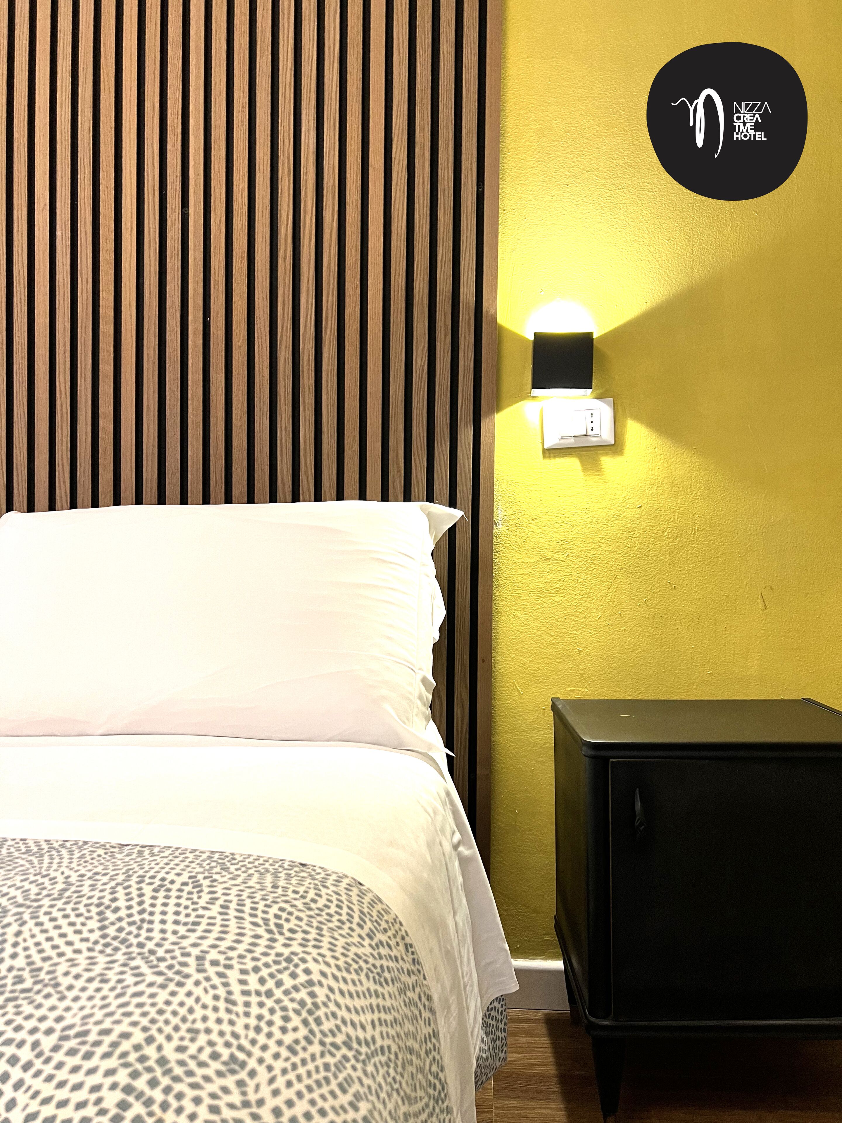 A hotel room with a bed, a nightstand, and a wall-mounted light fixture.
