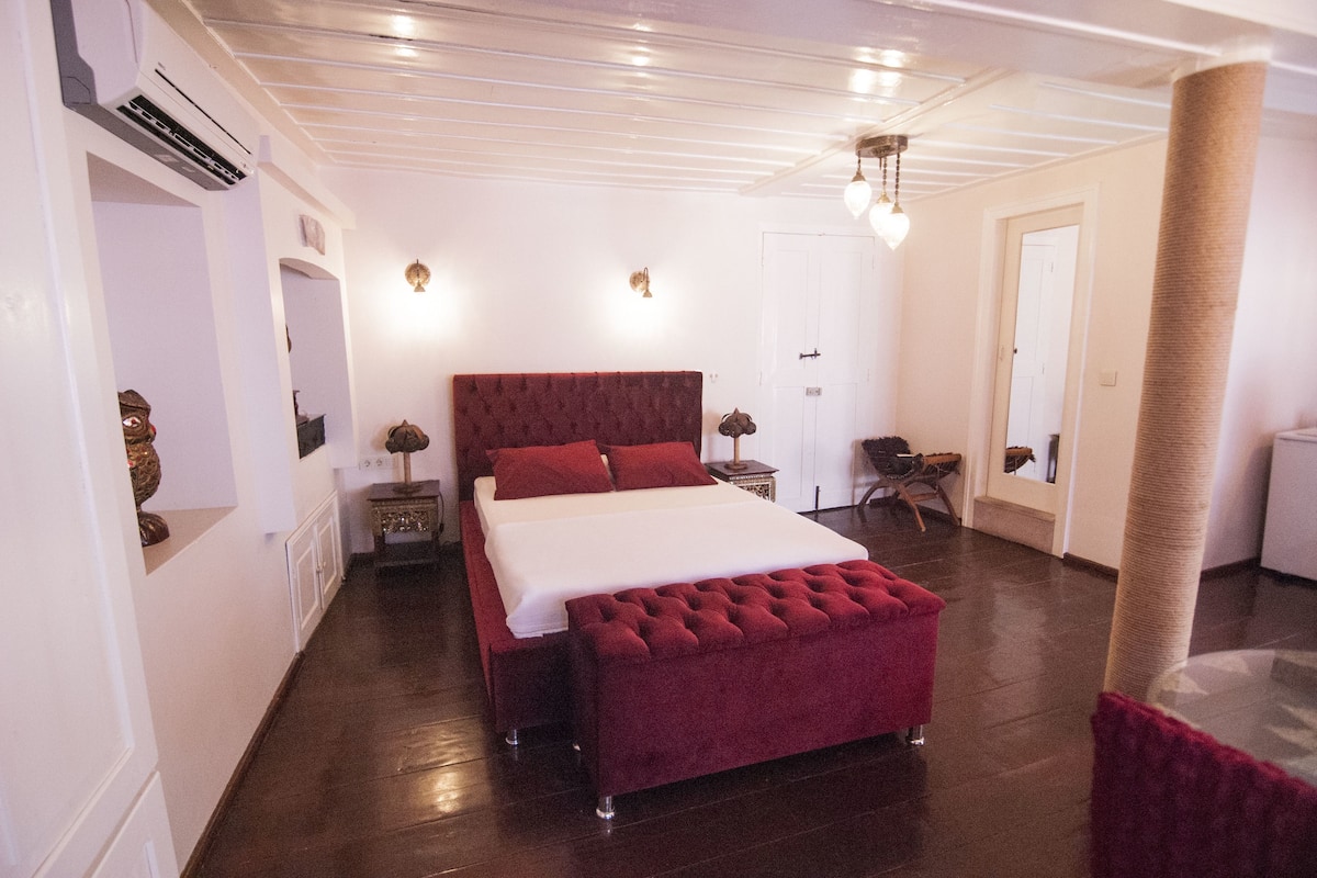 Um quarto espaçoso com uma cama grande, um banquinho estofado em vermelho, um arranhador para gatos e uma porta branca.