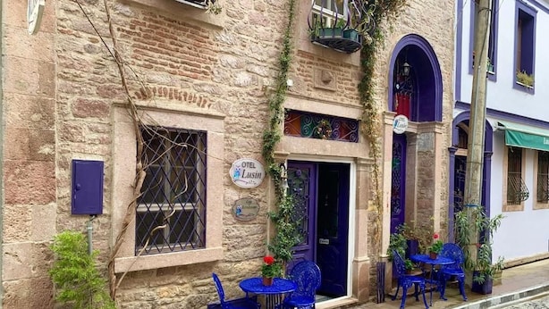 A stone building with a sign for 'OTEL Lusin' and a blue door. There are blue chairs and tables outside, and potted plants are visible. - Lusin Butik Hotel (Ayvalik)