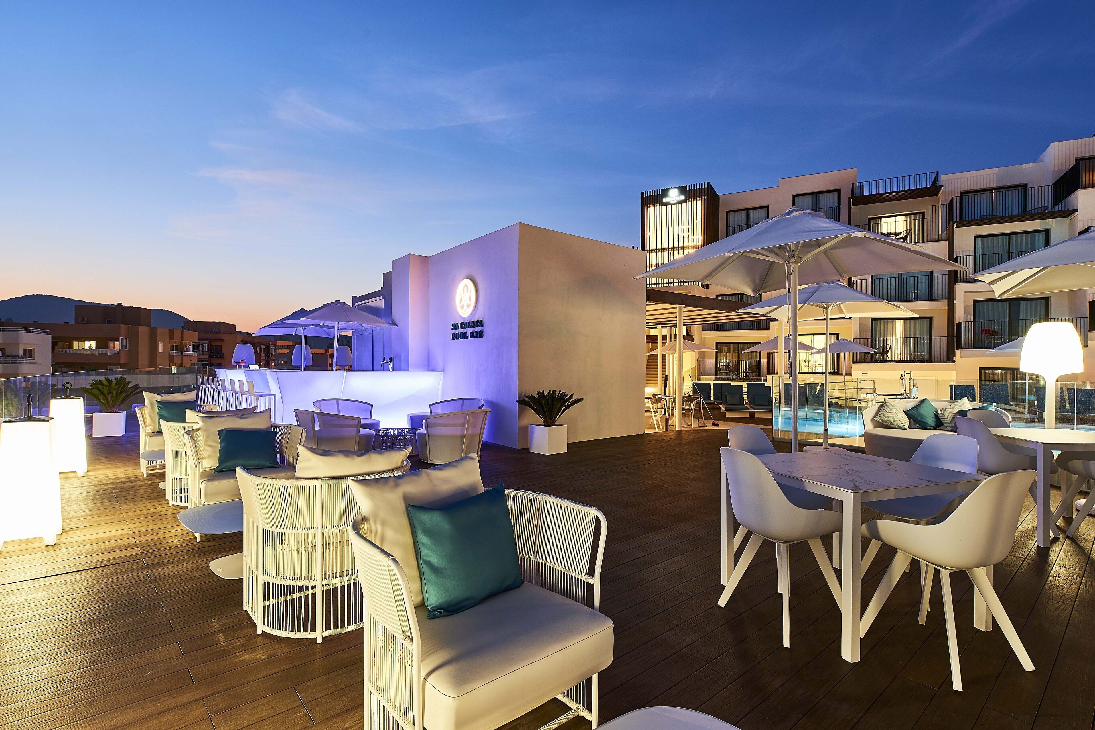 Un bar en la azotea con mesas al aire libre, piscina y un edificio moderno al fondo.