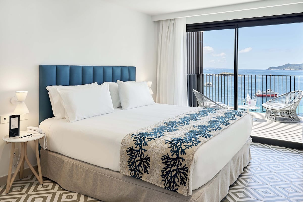 Een hotelkamer met een groot bed, een nachtkastje met telefoon, uitzicht op zee en een balkon met stoelen.