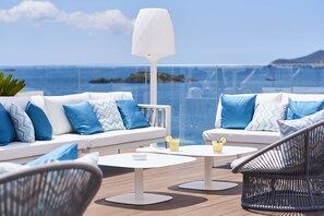 Un balcón con un sofá blanco, sillas de mimbre, una mesa blanca y barandilla de vidrio con vista al mar.