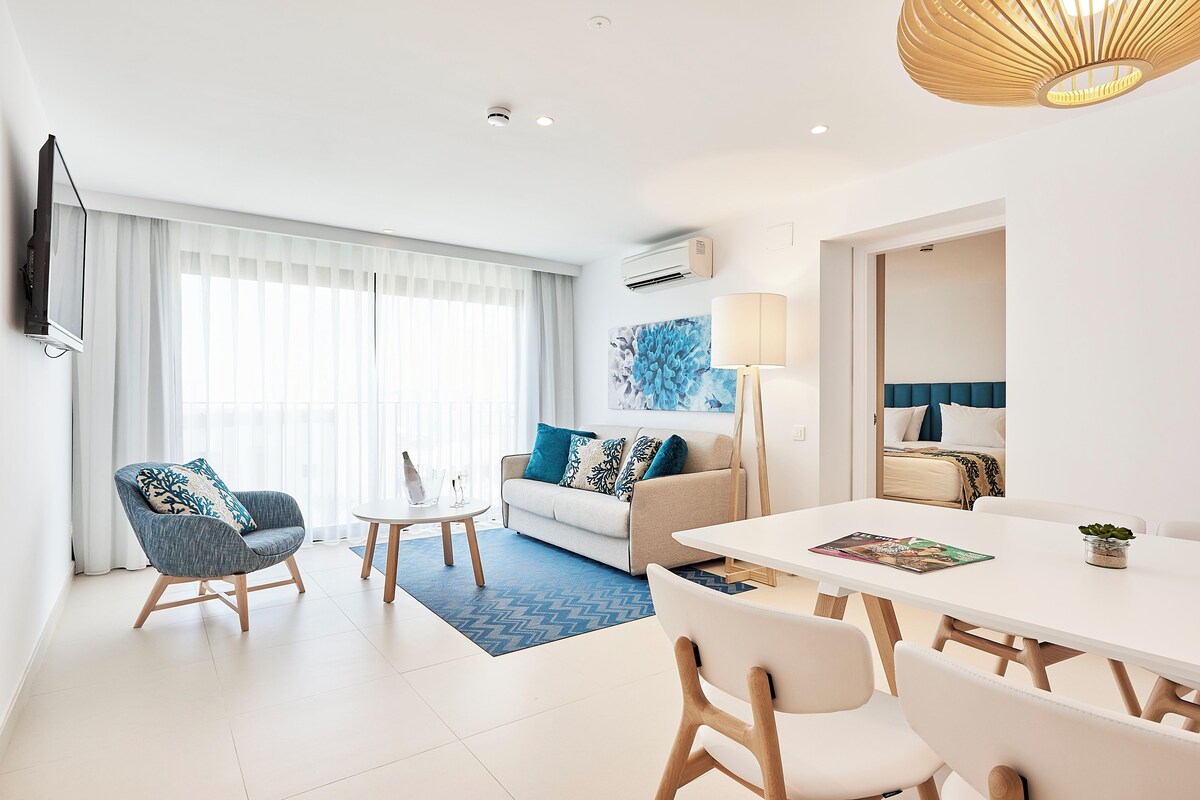 A modern living room with a sofa, armchair, dining table, and a TV mounted on the wall.