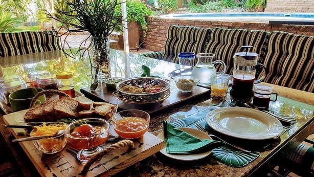 A breakfast setup with a variety of dishes, drinks, and a coffee pot on a glass table outdoors. - 800 at Merton (Pretoria)