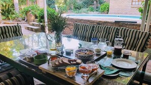 A dining table set with various dishes and drinks, surrounded by striped cushioned seating, with a pool visible in the background. - 800 at Merton (Pretoria)
