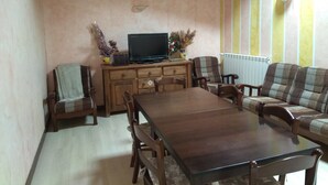 A cozy living room with a wooden dining table, two armchairs, a sofa, a television on a wooden cabinet, and a radiator. - La Cerca de Doña Jimena (Ibeas de Juarros)